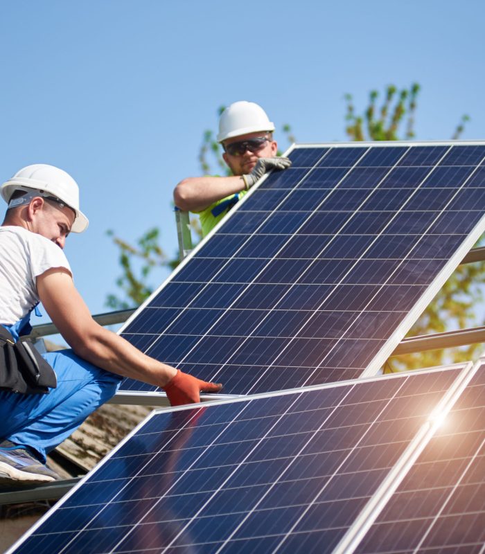 Two professional technicians adjusting heavy solar photo voltaic panel to metal platform on blue sky background. Stand-alone solar panel system installation, efficiency and professionalism concept.